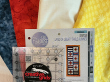 Load image into Gallery viewer, A packaged quilting set from Cut Loose Fabrics, featuring a 6 1/2 x 6 1/2 inch Creative Grids ruler and a pattern 'Land of Liberty Table Runner,' displayed over several fabric swatches.