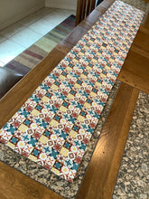 Load image into Gallery viewer, A reversible table runner with a Christmas-themed pattern featuring a variety of holiday motifs such as stars and Christmas trees, primarily in red, blue, and yellow hues, with metallic gold accents.