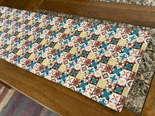 Load image into Gallery viewer, A reversible table runner with a Christmas-themed pattern featuring a variety of holiday motifs such as stars and Christmas trees, primarily in red, blue, and yellow hues, with metallic gold accents.