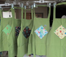 Load image into Gallery viewer, Five light green aprons hanging on a metal rack in a home setting, each featuring a different colorful diamond-shaped patchwork pocket.