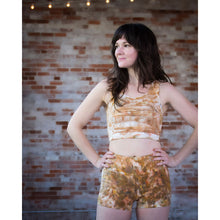 Load image into Gallery viewer, A woman modeling a matching activewear set consisting of a tie-dye crop top and high-waisted shorts in a warm brown, beige, and white pattern, standing against a brick wall background.

